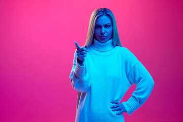 Irritated girl, young woman looking aggressively at camera and points her finger against gradient pink background in blue neon light.