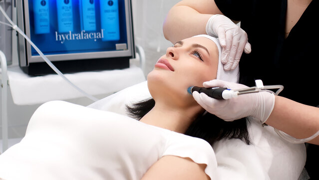 Cosmetology clinic. Professional female cosmetologist doing hydrafacial procedure while being a work. Attractive nice woman lying on the medical bed while having beauty procedures