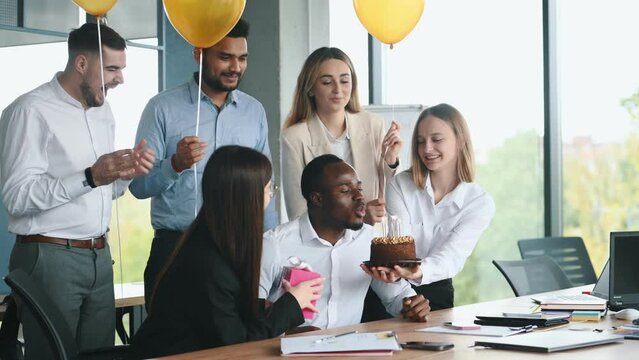 Young african american man is working in office then getting birthday cake from colleagues making wish blowing candle enjoying surprise having fun with creative team.