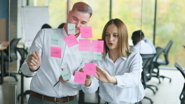 Video of successful business couple team writing on stickers at office glass board while discussing together in a modern office