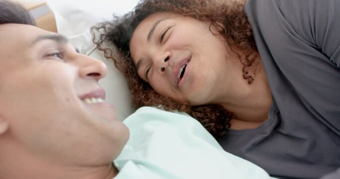 Happy diverse gay male couple lying on bed smiling and talking at home, slow motion