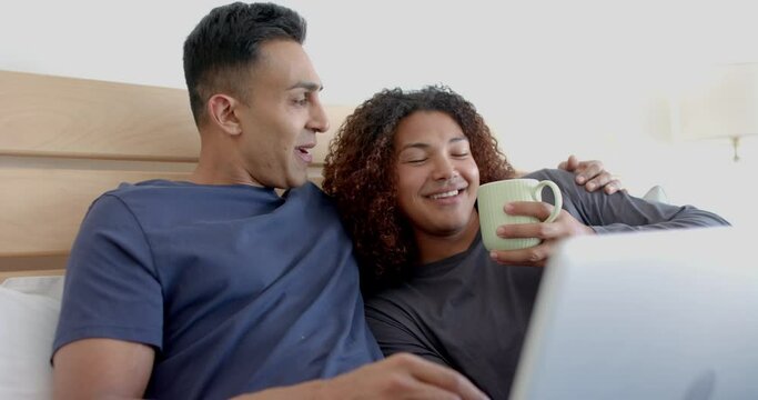 Happy diverse gay male couple on bed, having coffee and using laptop in the morning, slow motion
