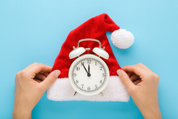 Woman hands holding bright red hat with white fur and alarm clock on light blue table background....