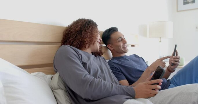 Happy diverse gay male couple sitting on bed using smartphones, talking in the morning, slow motion