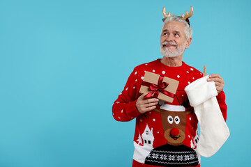 Senior man in Christmas sweater, reindeer headband holding gift and stocking on light blue background. Space for text