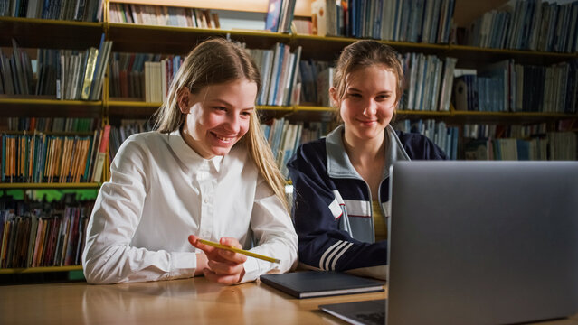 Two girl students sitting in the library and using a laptop during virtual online lessons. Internet, education, and e-learning concept.