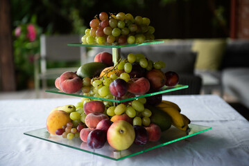 A three-tiered glass fruit bowl with many fresh fruits on a table with a white tablecloth - grapes, plums, bananas, peaches..