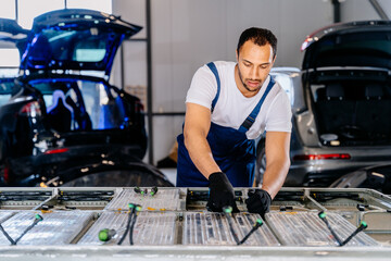 Portrait of biracial male expert technician repair electric car, EV car while opened. A used Lithium-ion car battery before its repair. Pack of battery cells module on platform.