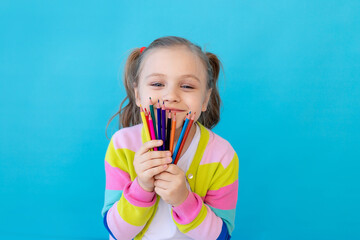 portrait of a cute little girl with colored pencils in a striped jacket. The concept of education and drawing. Photo studio, blue background, place for text
