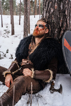 A Viking warrior with war paint on his face rests against a tree in the snow, appearing wounded and exhausted