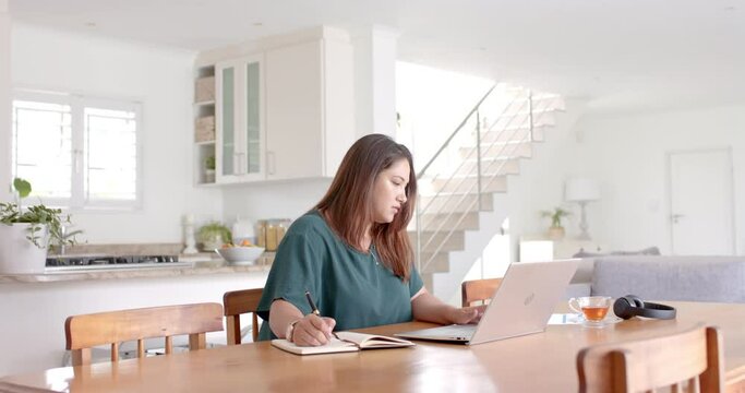 Plus size biracial woman making notes and using laptop, working from home, copy space, slow motion