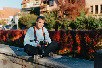 Obraz premium Man Sitting in Old European City And Holding Photo Camera. Contemporary Stylish Blogger And Photographer. Handsome man taking a selfie on a trip in Europe.