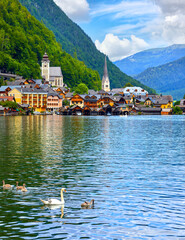 Hallstatt, Austria. View at Hallstattersee Lake and Alps mountains summits. White swan birds near the dock. Ancient houses lake banks with chapel. Summer day. Blue sky clouds