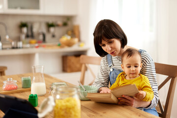 Young mother reading a book to her baby boy while having having breakfast in the dining room