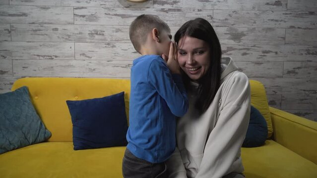 Medium shot. Cute son tells his mother secret, whispers in her ear, gossips about his personal life in the room, mom smiles, then hugs he son. spends free time together at home
