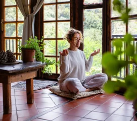 Abwaschbare Fototapete Full length attentive young Caucasian woman making mudra gesture, sitting in lotus position sitting on soft cushion on floor at home. Peaceful woman meditating deeply, doing breathing yoga  © simona