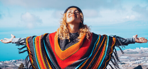 Blonde woman in hat and colorful poncho relaxing with open arms in blue winter sky. People's freedom style. - obrazy, fototapety, plakaty
