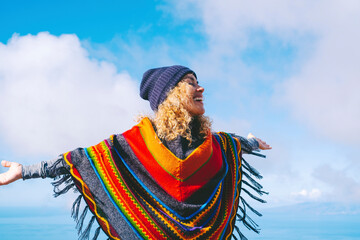 Blonde woman with hat and colorful poncho smiles and relaxes with open arms in the blue winter sky....