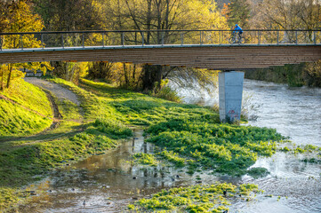 Holzbrücke mit Betonpfeilern über Fluss und Uferweg mit herbstlichen Bäumen
