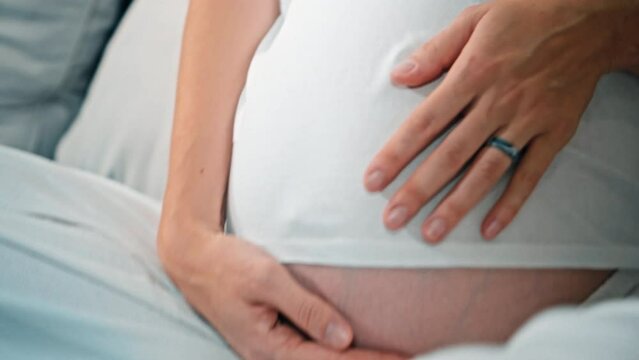 A young pregnant girl with a large stomach, sits at home on the sofa and touches her stomach with her hands.