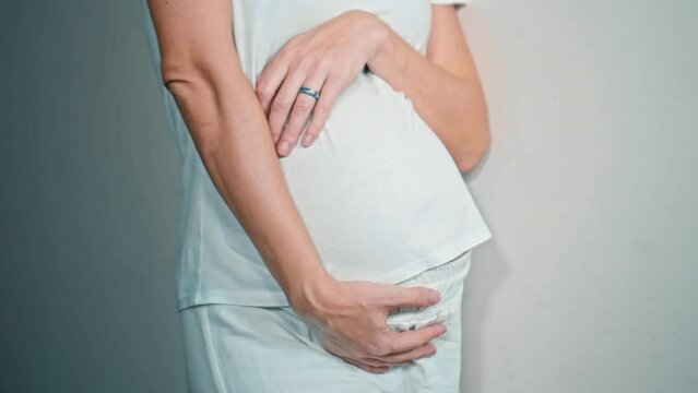 A young pregnant girl with a large stomach, stands at home against a white wall and touches her stomach with her hands.