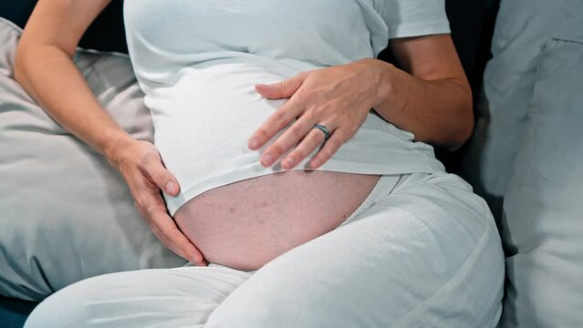 A young pregnant girl with a large stomach, sits at home on the sofa and touches her stomach with her hands.