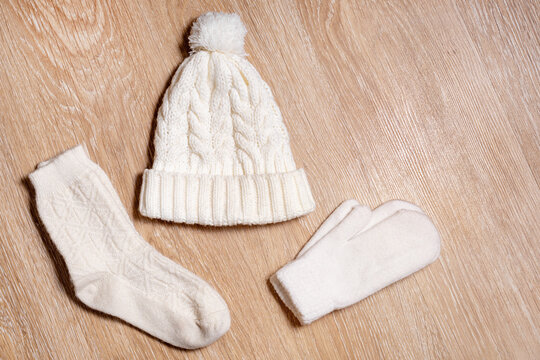 A white knitted hat with a pompom, woolen mittens and socks lie on wooden background. Details of winter clothing