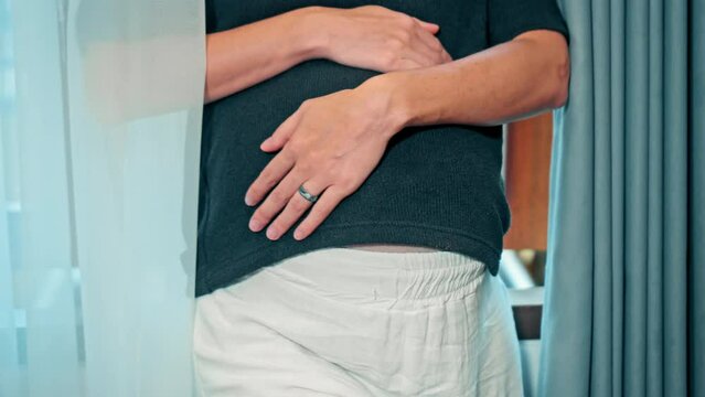 A young pregnant girl with a large stomach, stands at home by the window and touches her stomach with her hands.