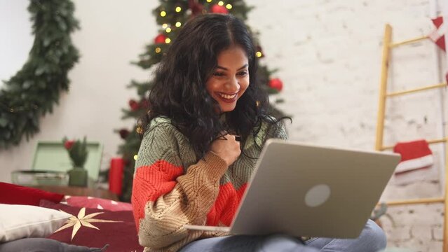Pretty young indian woman sitting on bed paying with credit card on laptop near Christmas decoration at cozy home Happy customer enjoying online shopping on holidays sales indoors
