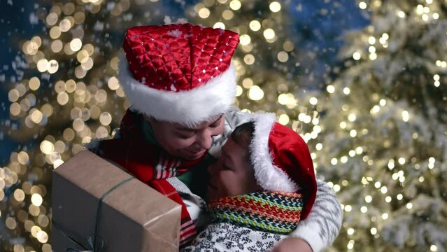 Smiling happy mom with xmas gift box and child in sweater with ornament enjoying winter holidays, blurred background of illuminated Christmas trees. High quality 4k footage