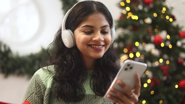 Charming young indian woman with headphones listening music while hold smartphone browsing scrolling app watching social media feed near Christmas tree with decoration at cozy home