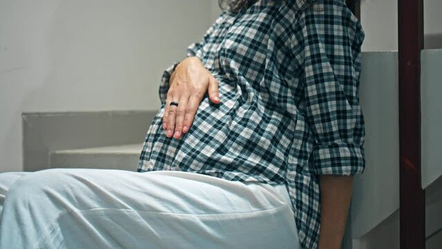 A young pregnant girl with a large stomach, stands at home on the stairs and touches her stomach with her hands.