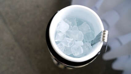 Ice in a blue transparent plastic bucket with ice cube tongs on a table background, top view. Blue...