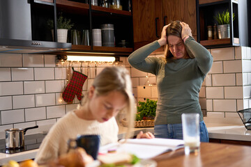 The daughter, looking weary, attempts to focus on her studies, while her stressed mother, holding her head, tries to convey the importance of perseverance. A challenging family scene in the kitchen.
