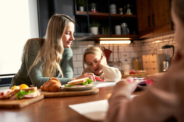 Mother and two daughters share the kitchen table, with mom and the elder sister working on homework together while the younger one joyfully doodles. A delightful morning of togetherness.