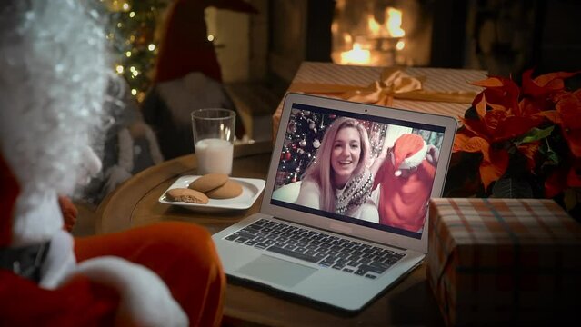 Santa Claus video calling family with little girl on laptop, greeting child by webcam, opening Christmas gift box in virtual online meeting using computer sitting in his decoration residence with