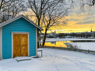 Bright blue wooden shed overlooking partially frozen lake covered in snow with golden orange sunset
