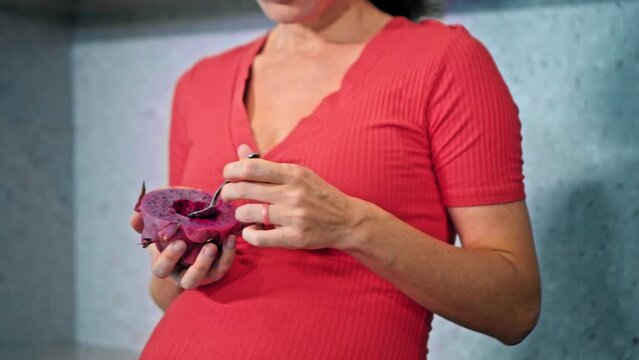 A young pregnant girl with a large stomach, stands at home in a colorful dress and eats dragonfruit.