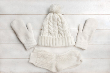 A white knitted hat with a pompom, woolen mittens and socks lie on a white wooden background....