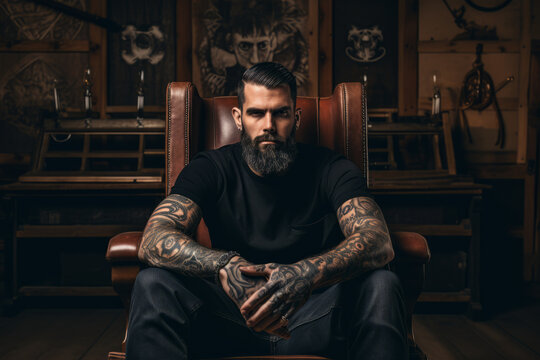 Handsome bearded man with tattooed arm sitting in leather chair and looking at camera
