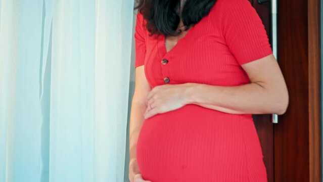 A young pregnant girl with a large stomach, stands at home in a colorful dress and touches her stomach with her hands.