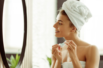 Smiling woman with towel on head applying patches, looking in mirror standing in stylish bathroom