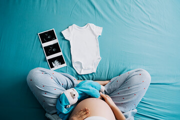 Woman pregnant belly with teddy toy bear, printed ultrasound images and baby white bodysuit. Concept of pregnancy, maternity, prepare for baby.