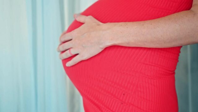 A young pregnant girl with a large stomach, stands at home in a colorful dress and touches her stomach with her hands.