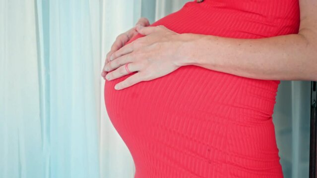 A young pregnant girl with a large stomach, stands at home in a colorful dress and touches her stomach with her hands.