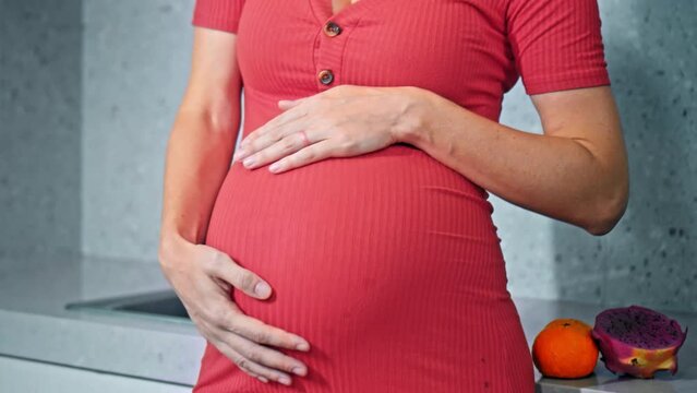 A young pregnant girl with a large stomach, stands in the kitchen in a colorful dress and touches her stomach with her hands.