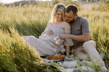 Happy pregnant couple spending time together on a picnic sitting outdoors at sunset in anticipation of a baby boy holding a shoe near the woman's belly.