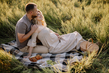 Naklejka na ściany i meble Beautiful pregnant couple in love spending time together in nature at sunset, having a picnic.