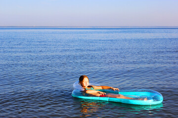 little happy girl in the sea with blond hair in a swimsuit floating in the sea on an inflatable blue mattress, Beautiful sunny seascape. the girl looks into the distance floats on calm water on the ho
