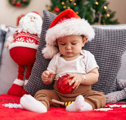Adorable hispanic baby holding christmas decoration ball sitting on sofa at home
